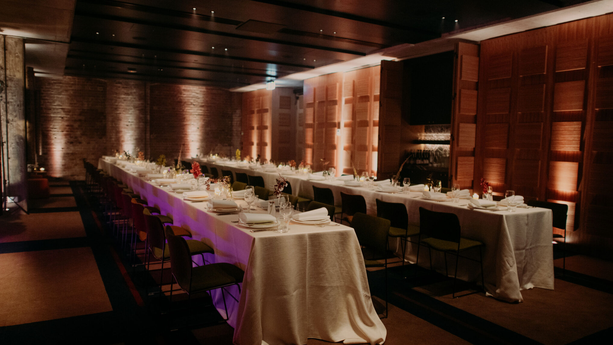 Two long banquet tables set for a formal dinner in a dimly lit, modern event space with brick and wood-paneled walls.